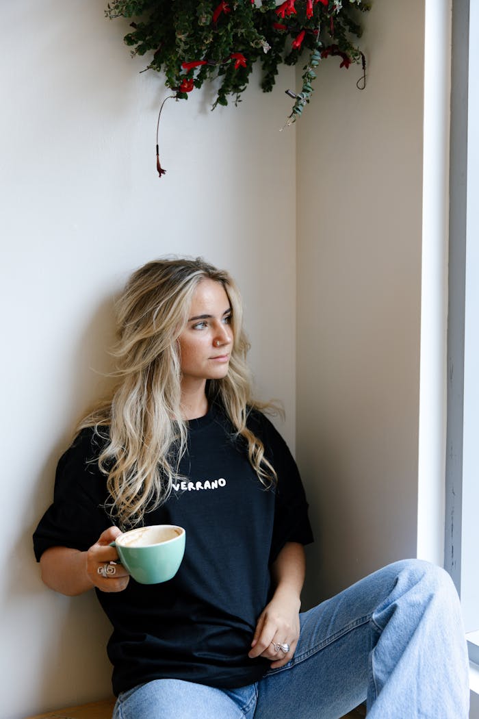 A young woman with long hair sitting by the window, holding a coffee cup indoors.